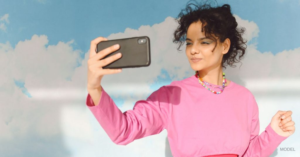 Woman holding a cell phone (model) shown taking a selfie in front of a background with painted clouds.